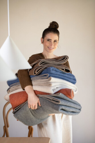 Junge Frau hält mehrere MERANO Kuscheldecken aus recycelter Wolle in natürlichen Farben – modernes, reduziertes Design